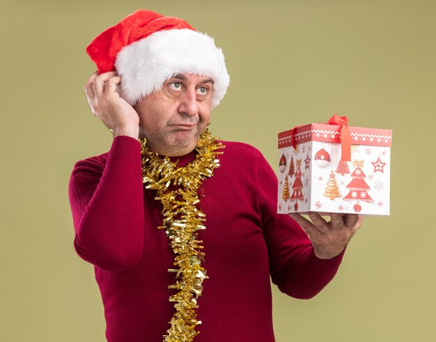 Hombre de mediana edad vestido con gorro de Papá Noel de Navidad con oropel alrededor del cuello sosteniendo un regalo de Navidad mirando hacia arriba desconcertado parado sobre fondo verde