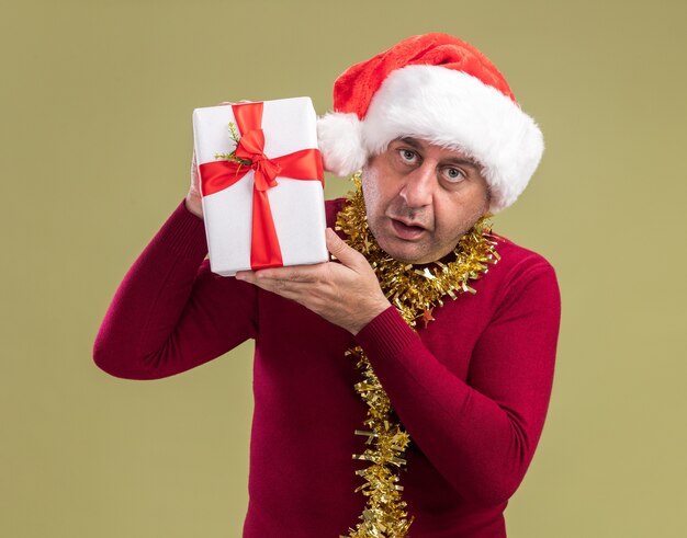 Hombre de mediana edad vestido con gorro de Papá Noel de Navidad con oropel alrededor del cuello sosteniendo el regalo de Navidad mirando a la cámara confundido de pie sobre fondo verde