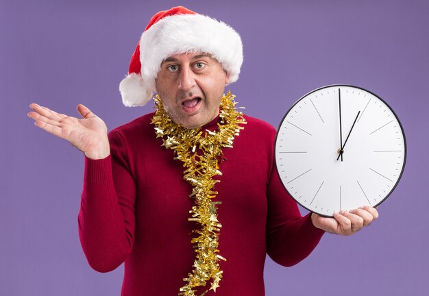 Hombre de mediana edad vestido con gorro de Papá Noel de Navidad con oropel alrededor del cuello sosteniendo el reloj mirando a la cámara confundido con el brazo levantado sobre fondo púrpura