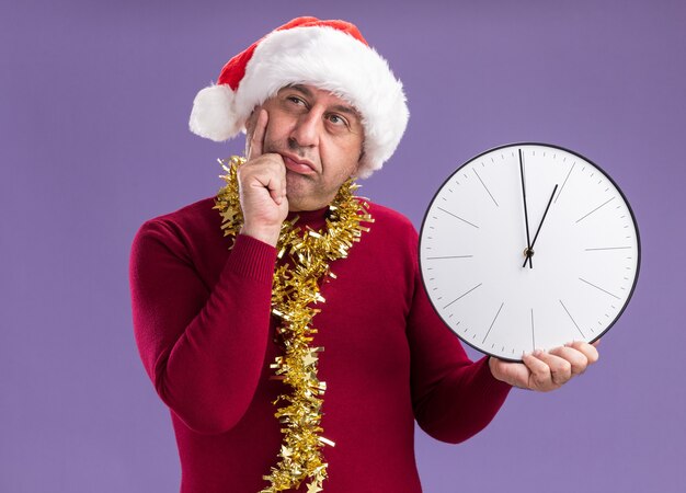 Hombre de mediana edad vestido con gorro de Papá Noel de Navidad con oropel alrededor del cuello sosteniendo el reloj mirando a un lado desconcertado de pie sobre fondo púrpura