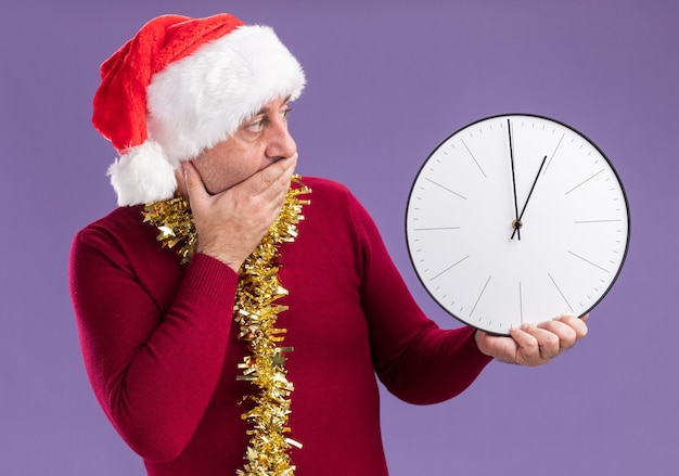 Foto gratuita hombre de mediana edad vestido con gorro de papá noel de navidad con oropel alrededor del cuello sosteniendo el reloj mirándolo preocupado cubriendo la boca con la mano de pie sobre fondo púrpura