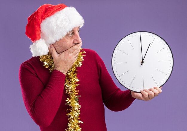 Hombre de mediana edad vestido con gorro de Papá Noel de Navidad con oropel alrededor del cuello sosteniendo el reloj mirándolo preocupado cubriendo la boca con la mano de pie sobre fondo púrpura