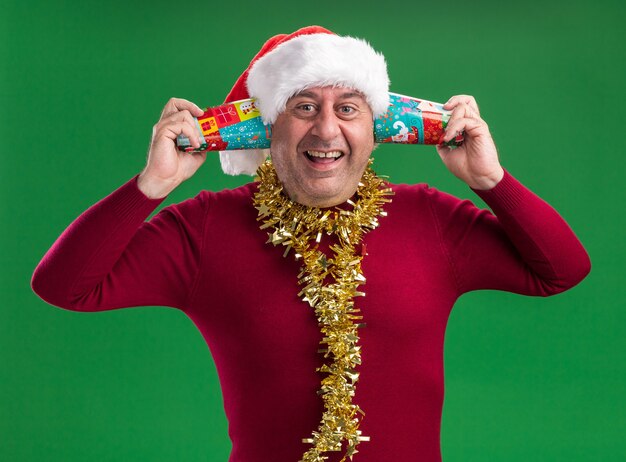 Hombre de mediana edad vestido con gorro de Papá Noel de Navidad con oropel alrededor del cuello sosteniendo vasos de papel de colores sobre su oreja, feliz y emocionado de pie sobre la pared verde