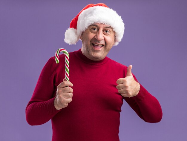 Hombre de mediana edad vestido con gorro de Papá Noel de Navidad sosteniendo bastón de caramelo con una sonrisa en la cara mostrando los pulgares para arriba sobre la pared púrpura