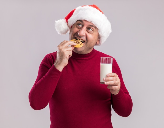 Hombre de mediana edad vestido con gorro de Papá Noel de Navidad sosteniendo un vaso de leche comiendo galletas sonriendo feliz y alegre de pie sobre la pared blanca