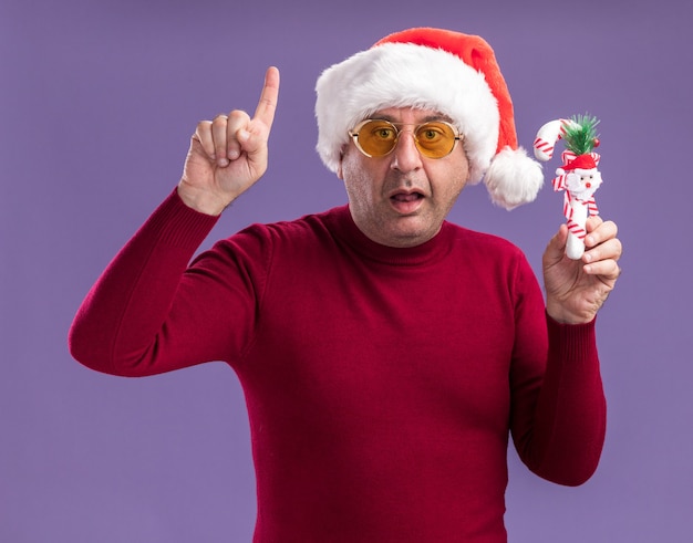 Hombre de mediana edad vestido con santa navidad en gafas amarillas sosteniendo bastón de caramelo de navidad mirando a la cámara sorprendido mostrando el dedo índice de pie sobre fondo púrpura