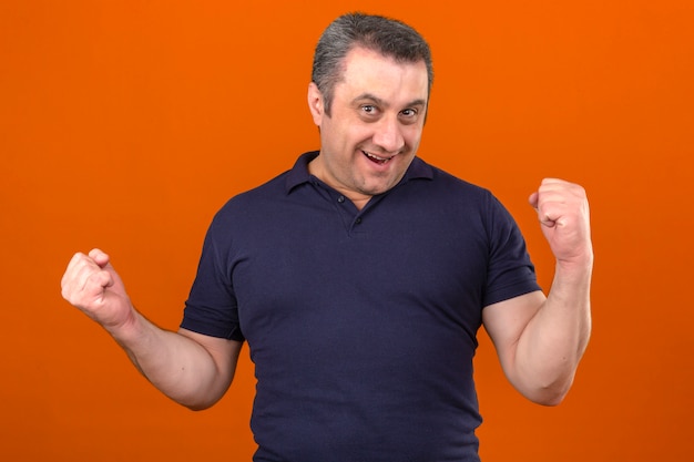 Hombre de mediana edad vistiendo camisa polo sonriendo levantando los puños como un ganador sobre la pared naranja aislada