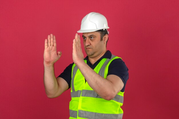 Hombre de mediana edad vistiendo chaleco amarillo de construcción y casco de seguridad dirigiendo gesticulando con las manos mostrando el tamaño sobre la pared rosa aislada