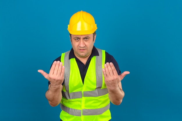 Hombre de mediana edad vistiendo chaleco amarillo de construcción y casco de seguridad guiando a alguien a la dirección sobre la pared azul aislada