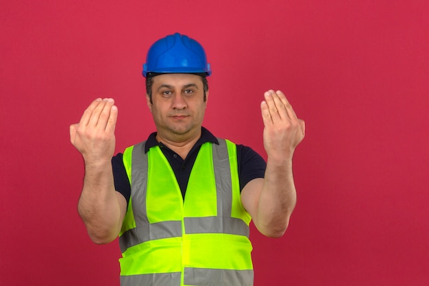 Foto gratuita hombre de mediana edad vistiendo construcción chaleco amarillo y casco de seguridad guiando a alguien a la dirección sobre pared rosa aislado