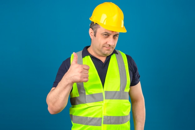 Hombre de mediana edad vistiendo construcción chaleco amarillo y casco de seguridad sonriendo y mostrando los pulgares hacia arriba sobre la pared azul aislada