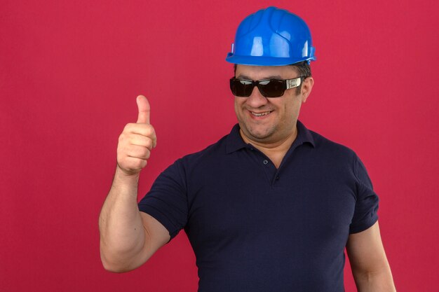 Hombre de mediana edad vistiendo polo y casco de seguridad mostrando los pulgares para arriba con una sonrisa en la cara sobre la pared rosa aislado