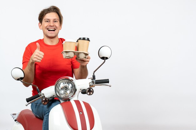 Hombre de mensajería sonriente joven en uniforme rojo sentado en scooter y manteniendo órdenes haciendo gesto ok en la pared amarilla