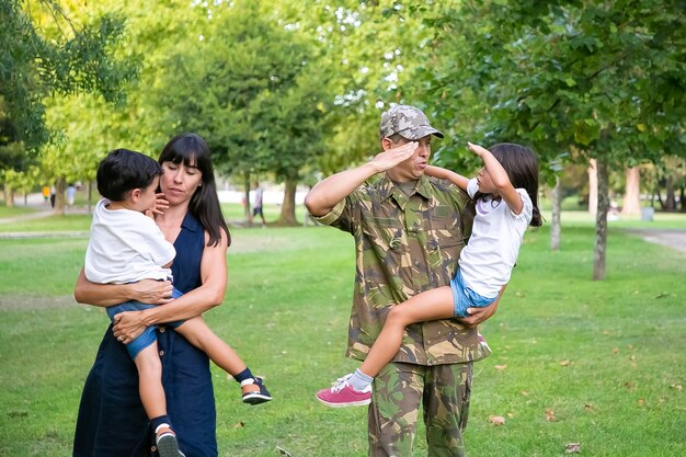 Hombre militar positivo caminando en el parque con su esposa e hijos, enseñando a su hija a hacer el gesto de saludo del ejército. Vista trasera completa. Reunión familiar o concepto de padre militar