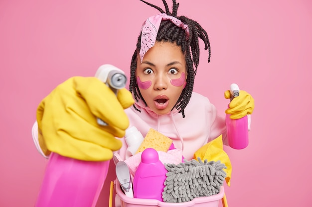 El hombre mira con expresión omg sostiene suministros de limpieza listos para limpiar la habitación se somete a procedimientos de belleza mientras posa en interiores en rosa
