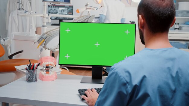 Hombre mirando la computadora con pantalla verde horizontal para odontología en el gabinete. Asistente trabajando con clave de croma y antecedentes aislados en el monitor para atención estomatológica y chequeo