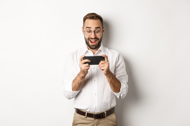 Hombre mirando emocionado y sorprendido por el teléfono móvil, sosteniendo el teléfono inteligente horizontalmente, de pie.