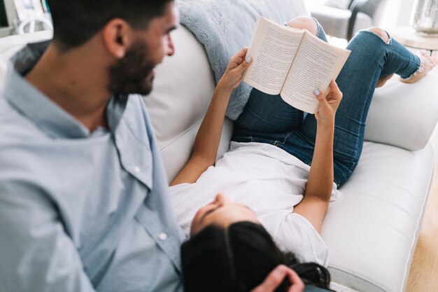 Hombre mirando a mujer tendida en el sofá sosteniendo el libro