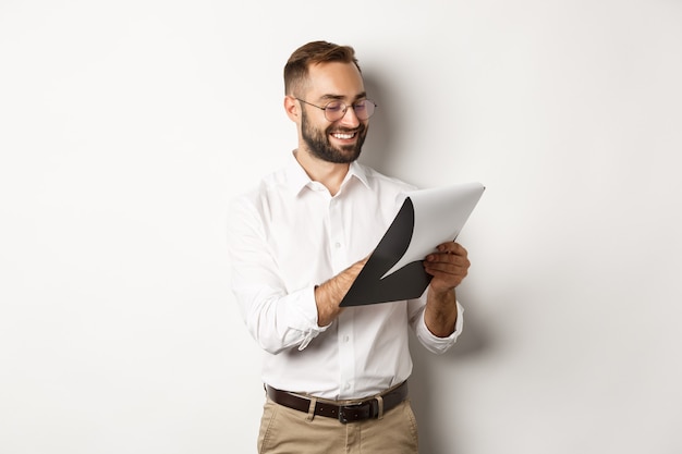 Hombre mirando satisfecho mientras lee documentos, sosteniendo el portapapeles y sonriendo, de pie