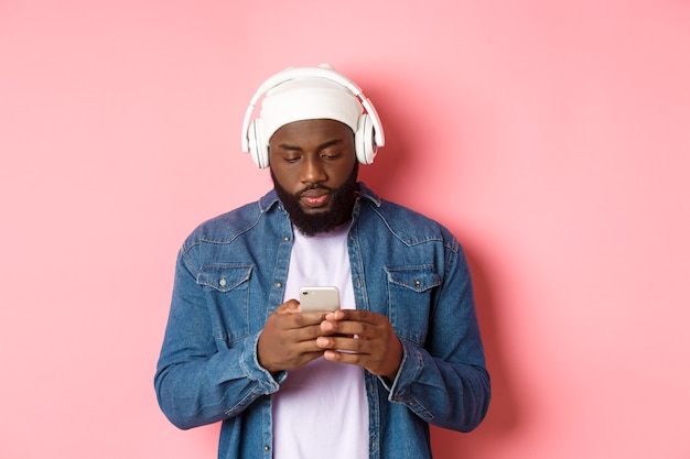 Hombre mirando serio mientras lee un mensaje en el teléfono, escuchando música en auriculares, de pie sobre un fondo rosa.