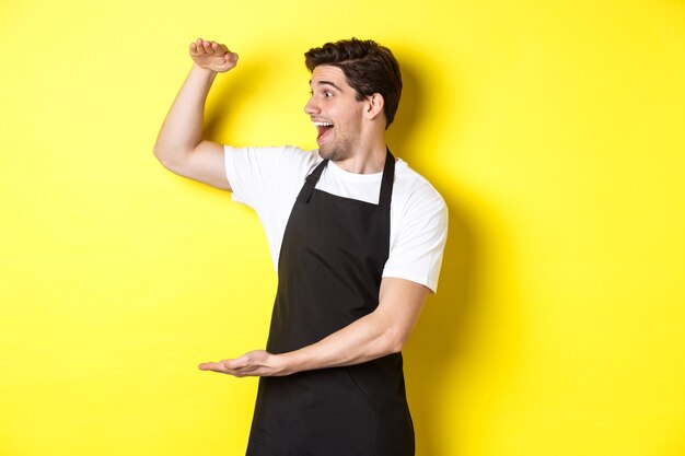 Hombre mirando sorprendido por algo grande, de pie con delantal negro sobre fondo amarillo.