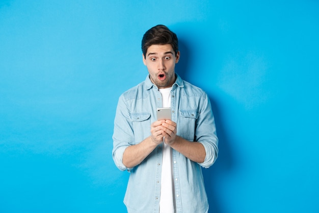 Hombre mirando sorprendido mientras revisa la promoción en el teléfono inteligente, mirando sorprendido por el teléfono, de pie contra el fondo azul.