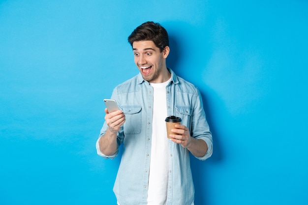 Hombre mirando sorprendido por el teléfono inteligente, tomando café, parado sobre fondo azul asombrado