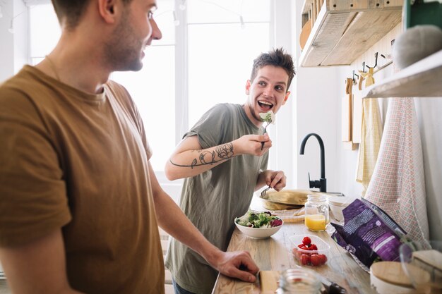 Hombre mirando a su amigo comiendo ensalada con un tenedor en la cocina