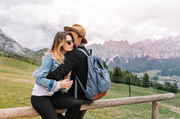 Hombre con mochila azul con elegante sombrero abrazando a su novia y disfrutando del increíble paisaje de montaña