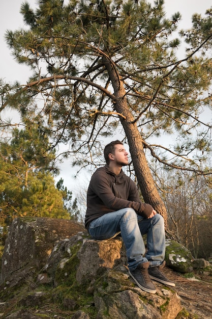 Hombre con mochila sentado en las rocas del bosque y mirando el horizonte en un día sombrío