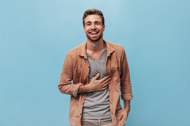Hombre de moda alegre con barba de jengibre en camiseta gris y camisa elegante riendo y posando sobre fondo azul aislado