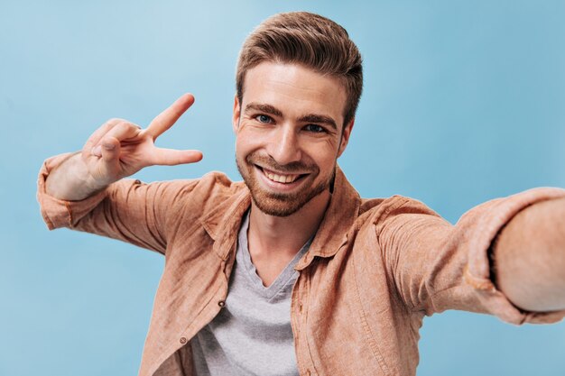 Hombre de moda con barba fresca en camiseta gris y camisa marrón haciendo fotos y mostrando el signo de la paz en la pared azul aislada