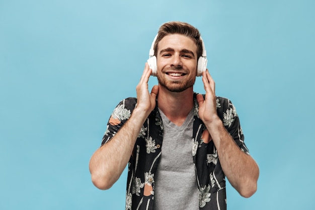 Hombre de moda sonriente con barba en camisa moderna y camiseta de planes escuchando música y mirando a la cámara sobre fondo azul