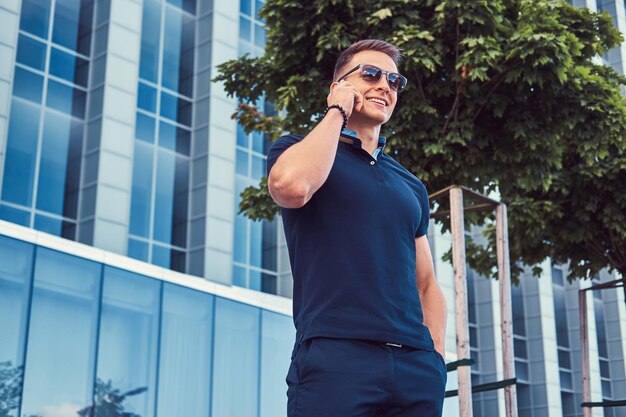 Un hombre de moda sonriente y guapo con un elegante corte de pelo con gafas de sol, vestido con una camiseta negra y pantalones, habla en el teléfono inteligente, se encuentra en la ciudad moderna contra un rascacielos.