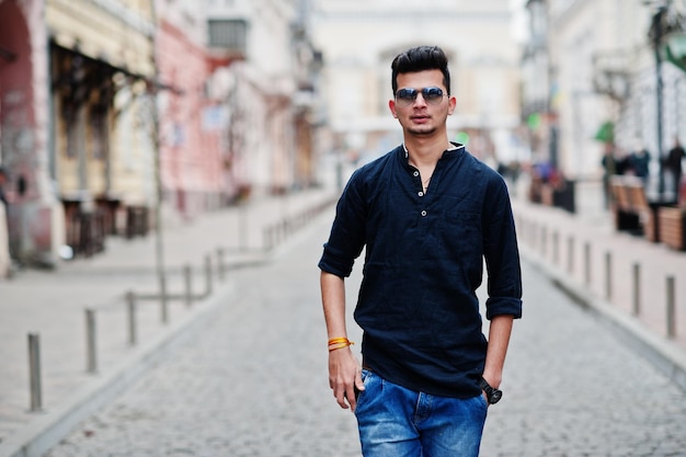Hombre modelo indio elegante con ropa informal, camisa negra y gafas de sol posadas al aire libre en la calle de la India