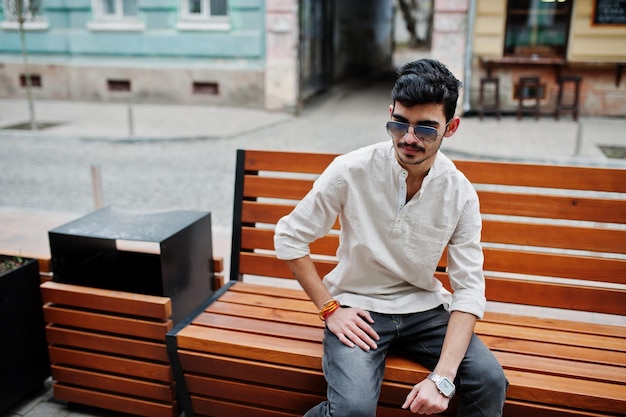 Hombre modelo indio elegante con ropa informal y gafas de sol posado al aire libre en la calle de la India y sentado en un banco