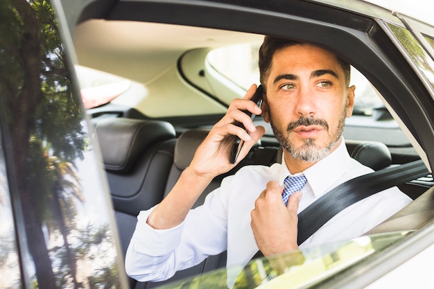 Hombre moderno que se sienta en el coche que ajusta su corbata que habla en el teléfono móvil