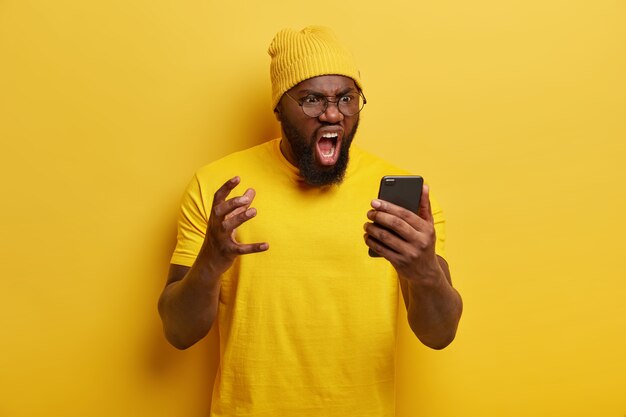 Hombre molesto con barba espesa grita enojado, tiene expresión facial furiosa, recibe noticias desagradables, usa un sombrero amarillo vivo y una camiseta