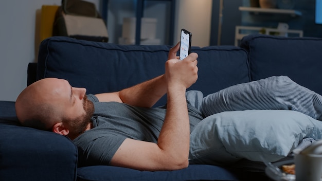 Hombre molesto causado por la ruptura acostado en el sofá en casa navegando en el teléfono inteligente