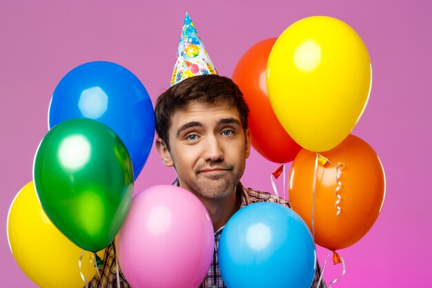 Hombre molesto celebrando cumpleaños, sosteniendo globos de colores sobre la pared púrpura.