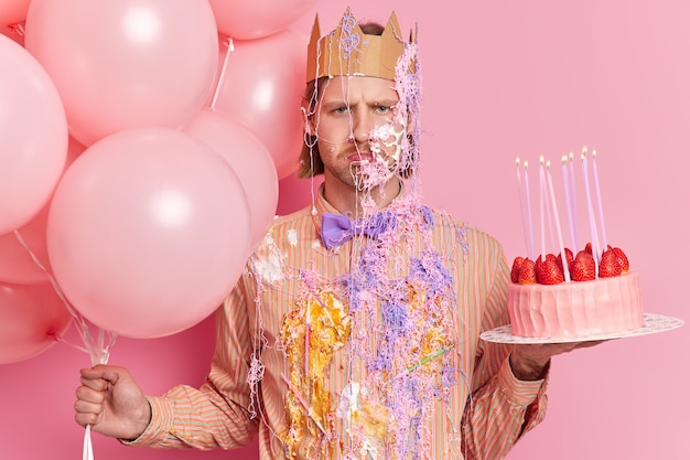 Hombre molesto sombrío decepcionado untado con crema y spray de serpentina listo para celebrar poses de cumpleaños con globos de aire y tarta de fresa