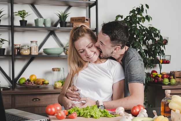 Hombre mordiendo en las mejillas de mujer de pie detrás del mostrador de la cocina con verduras