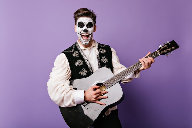 Hombre morena emocionado con maquillaje zombie cantando en la pared púrpura. Filmación en interiores de muerte chico tocando la guitarra y riendo.