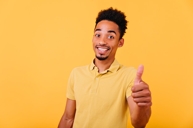 Hombre morena extasiado en traje de verano brillante posando con una sonrisa. Filmación en interiores de alegre modelo africano mostrando el pulgar hacia arriba y riendo.