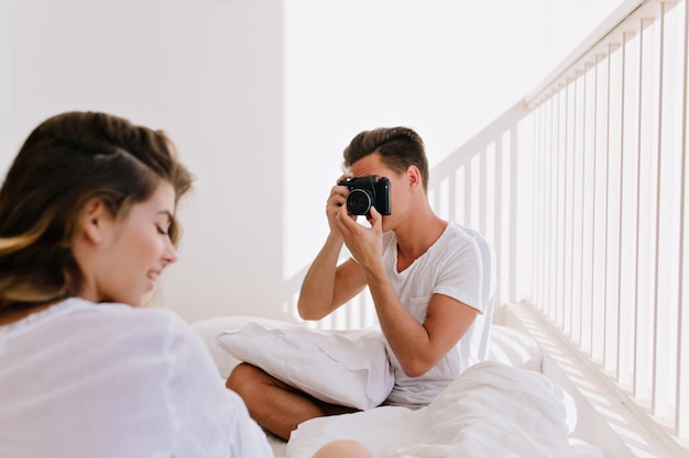 Hombre morena con peinado de moda haciendo foto de su hermosa novia, mientras descansa en fin de semana en el balcón. Agraciada mujer joven con camisa blanca posando para su novio fotógrafo