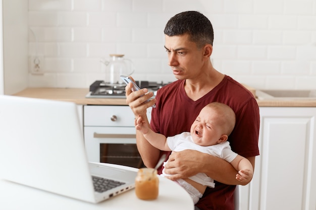 Hombre morena sorprendido con camiseta marrón estilo casual sentado a la mesa en la cocina con su hija pequeña, mirando la pantalla del teléfono inteligente con expresión asustada.