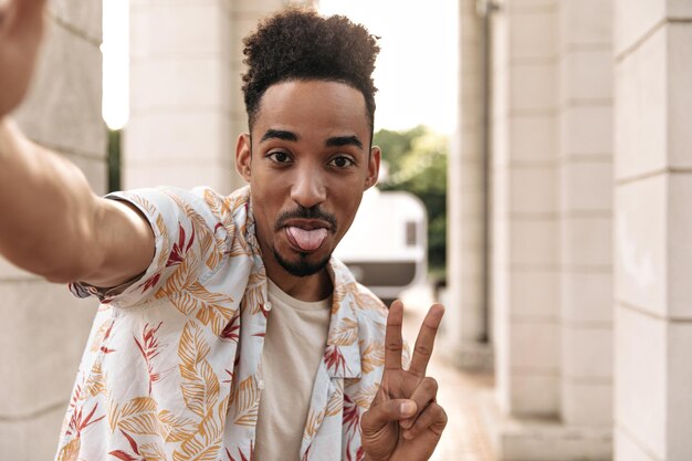 Hombre moreno rizado barbudo de ojos marrones con camiseta beige y camisa floral muestra el signo de la paz de la lengua y se toma una selfie afuera