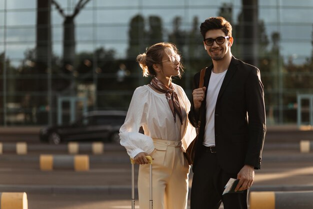 Hombre moreno con traje negro y gafas de sol sonríe Atractiva mujer rubia con blusa blanca mira a su novio Los viajeros de negocios posan cerca del aeropuerto