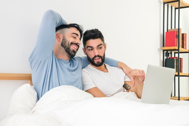 Hombre mostrando algo en la computadora portátil a su novio sentado en la cama