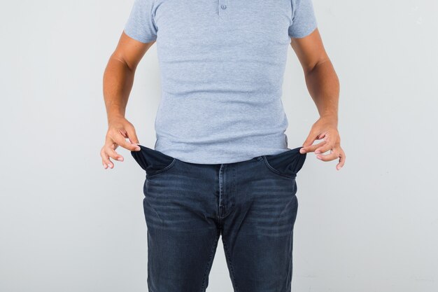 Hombre mostrando bolsillos vacíos en camiseta gris, jeans